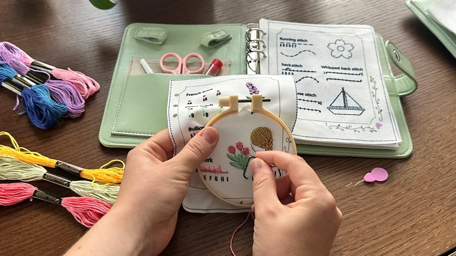 Hands stitching a hooped linen page in front of the open Embroidery Stitch Book and colorful floss